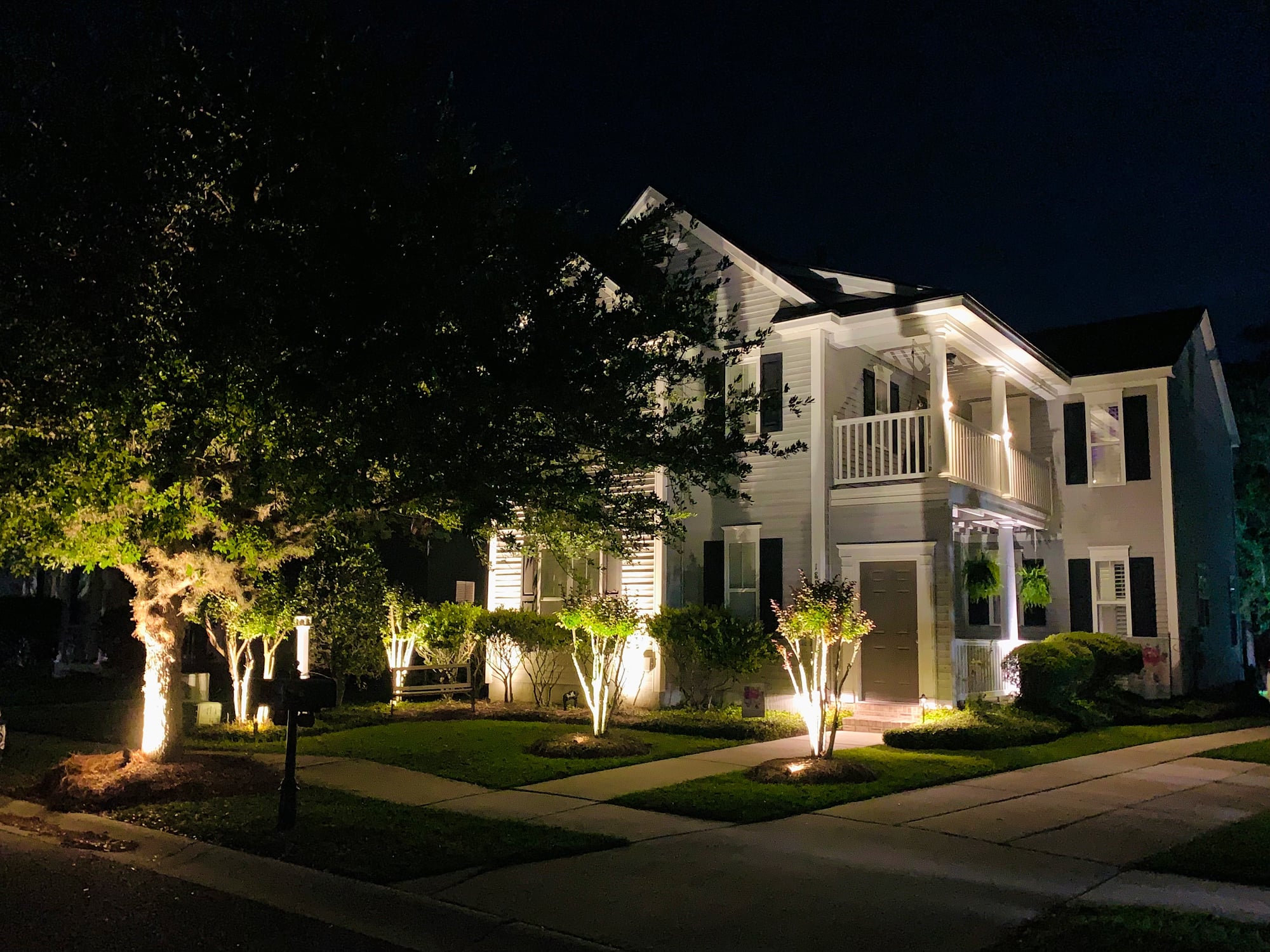 Night exterior of a white two-story home with black shutters, lit by warm landscape uplights on ornamental trees and shrubs across the front yard, with porch string lights and a lit covered entryway.