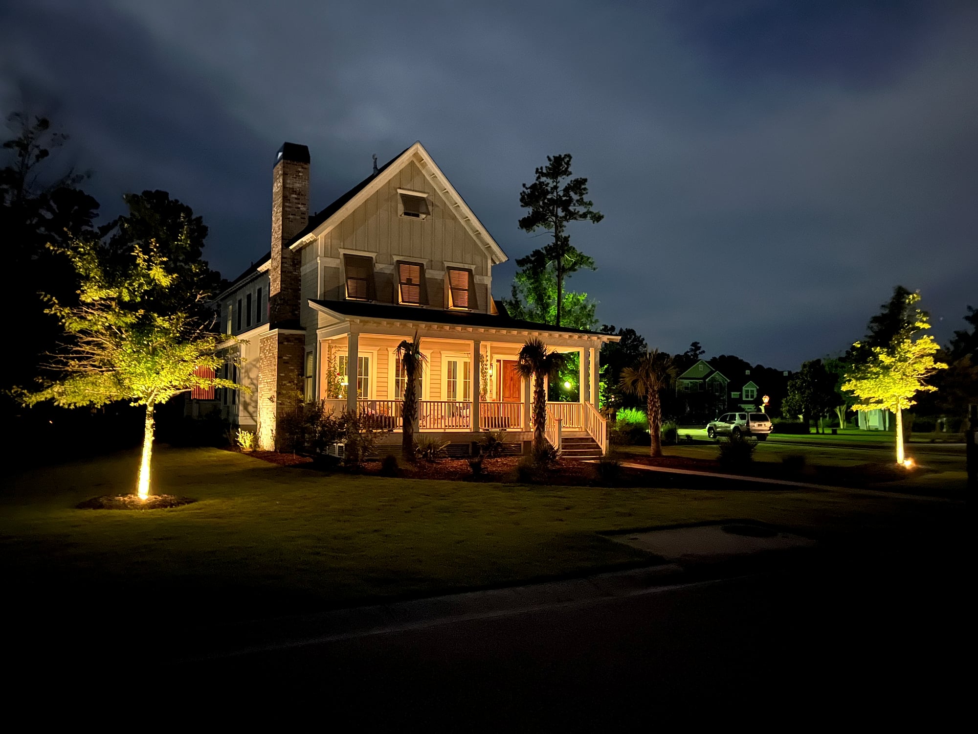 Nighttime landscape lighting on a two-story Southern farmhouse-style home, with warm yellow-green uplights illuminating flanking trees, soft porch lighting, and a deep blue dusk sky in the background.