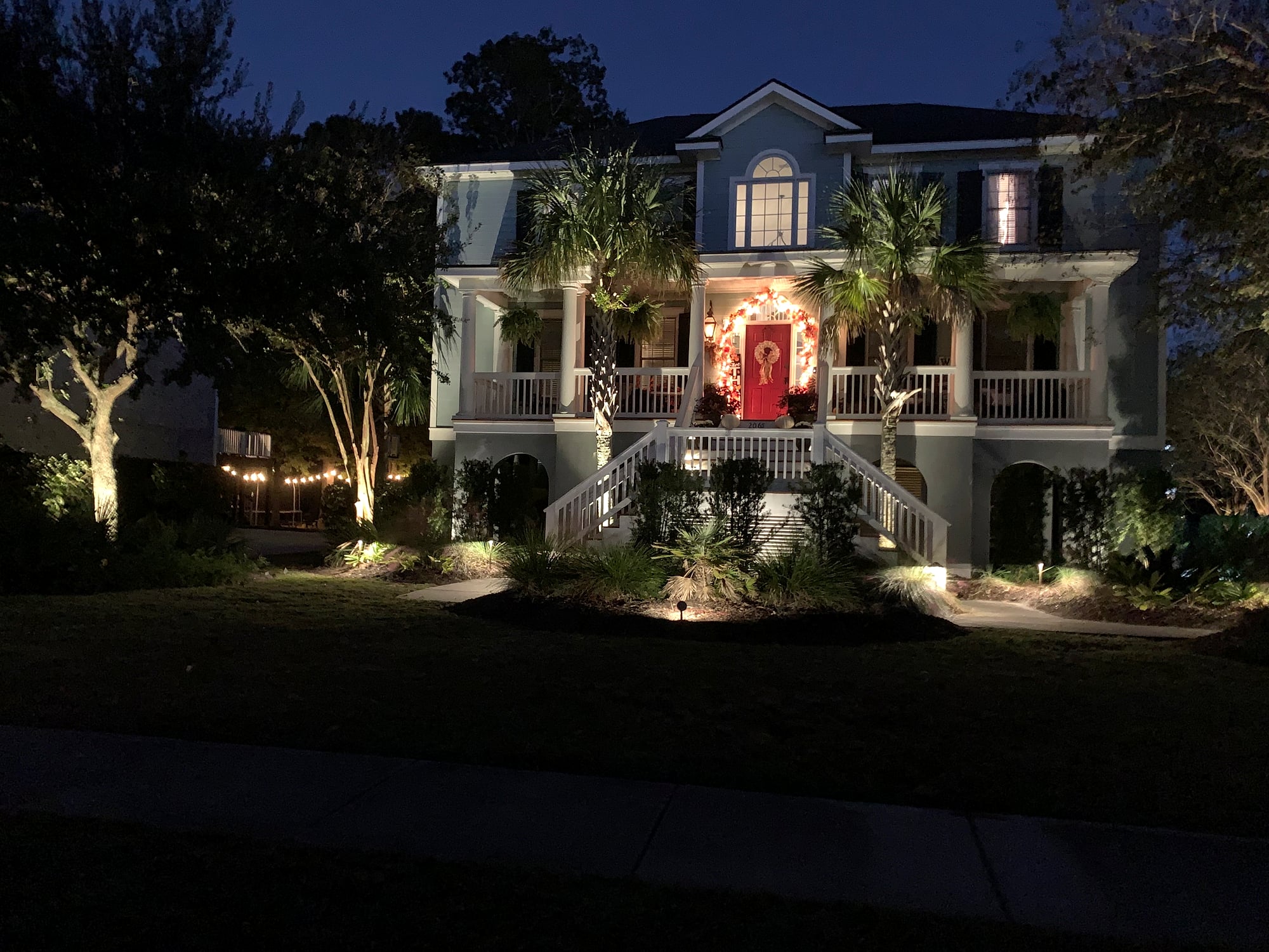 Night exterior of a two-story light-blue Charleston-style home with professional landscape uplighting illuminating palm trees and oaks, ground-level path lights lining the garden beds, and a glowing red front door framed by a holiday wreath, set against a deep blue evening sky.