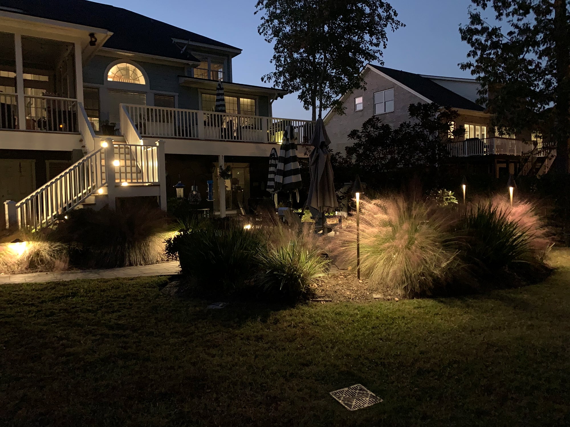 Dusk backyard of a two-story coastal home with warm amber path lights illuminating ornamental grasses, stair step lighting on a white-railed deck, and uplighting along a mulched garden bed under a deep blue twilight sky.