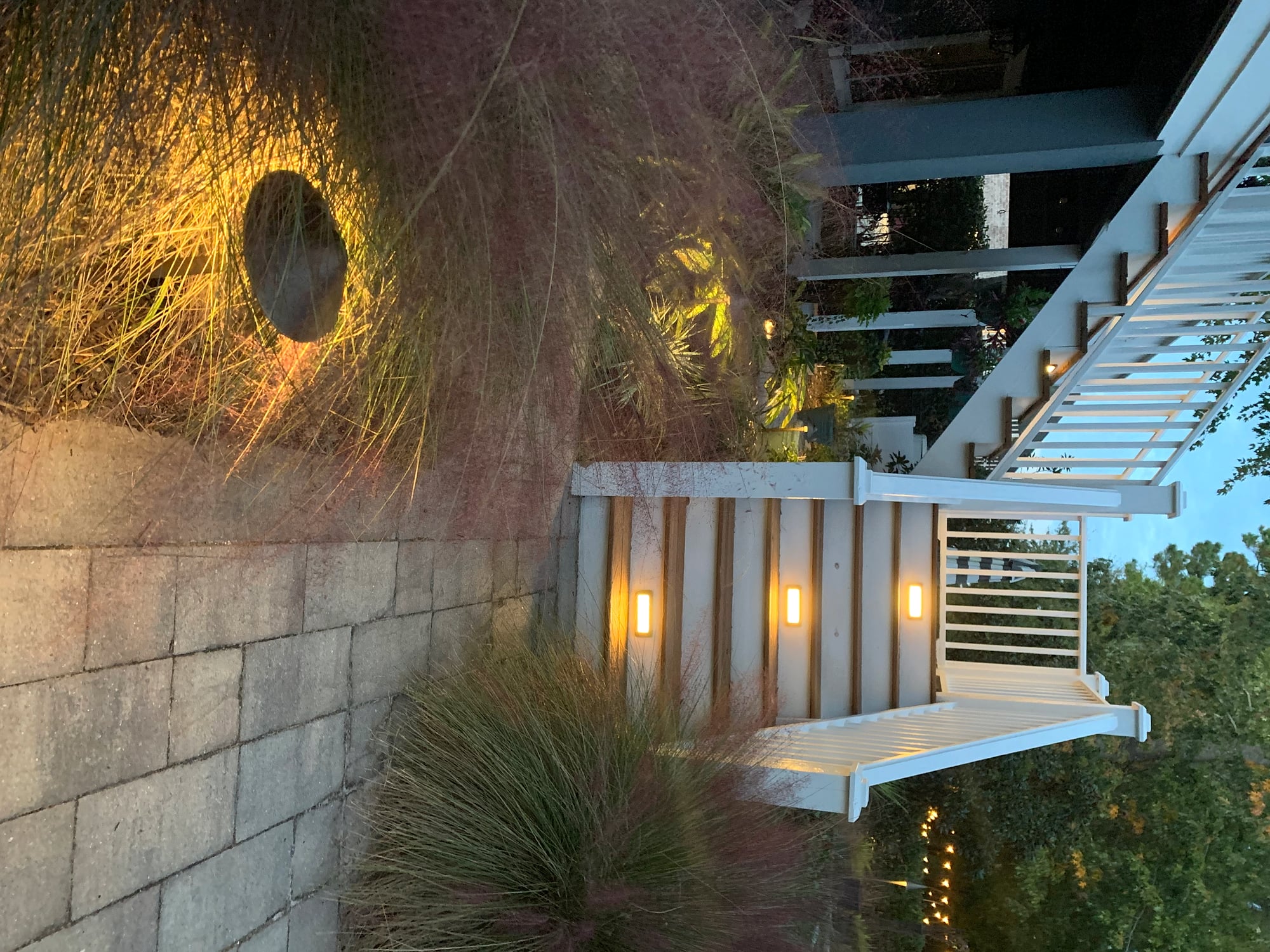 Dusk landscape lighting scene featuring two warm-white rectangular wall sconces mounted on a white deck fascia and a single ground uplighter casting a golden glow through tall ornamental grasses along a concrete block retaining wall, with a staircase descending into a garden bed.